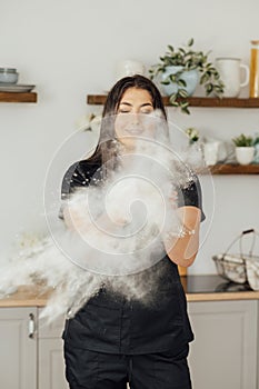 Woman baker clapping and sprinkling white flour over dough on kitchen background