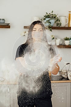 Woman baker clapping and sprinkling white flour over dough on kitchen background