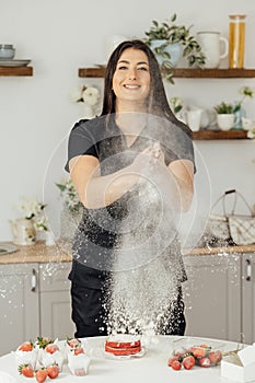 Woman baker clapping and sprinkling white flour over dough on kitchen background