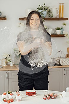 Woman baker clapping and sprinkling white flour over dough on kitchen background