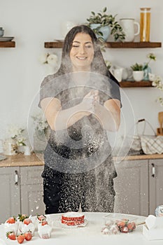 Woman baker clapping and sprinkling white flour over dough on kitchen background