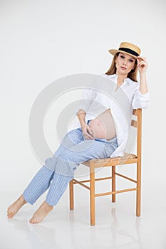 Woman with bare belly in late pregnancy sit on chair holding hat on head, white background. Portrait of young pregnant