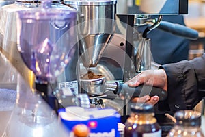 A woman barista spooning a coffee powder from the modern stainless steel grinder with a demitasse spoon at the coffee shop kitchen