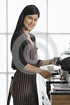 Woman barista wearing an apron standing in front of coffee machine and making beverage in coffee shop and looking to camera with