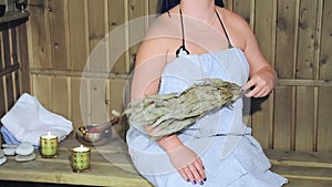 A woman in a bath in a towel without a face fanns herself with a fragrant laurel broom