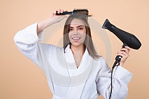 Woman in bathrobe combing hair, drying hairs with hairdryer in studio. Portrait of female model with a comb brushing