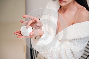 Woman in bathrobe holding jar with face cream, applying moisturizing product on skin
