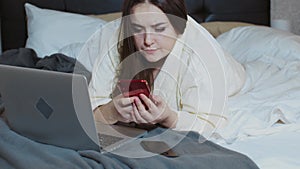 Woman in a bathrobe lying on the bed using a laptop and smartphones for work. Freelance, work at home, work outside the