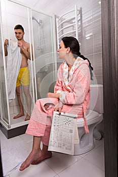 Woman in bathrobe with newspaper sitting on a toilet and waiting