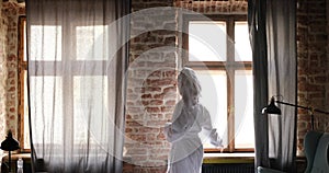 Woman in bathrobe opening curtain and looking out through window