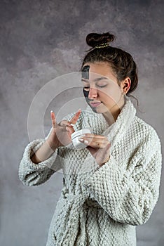 woman in bathrobe relaxing with black face mask and holding jar with cream isolated on grey