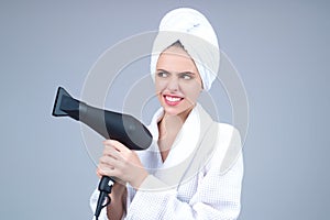 Woman in bathrobe and towel on head with hairdryer in studio. Portrait of female model with blow dryer. Hair care and