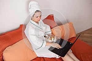 A woman in a bathrobe and a towel on her head at home on a bed with a cat and a laptop looks into the computer monitor.