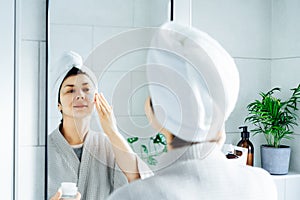 Woman in bathrobe with a towel on her head looking in the mirror and applying moisturizer cream to her face in the