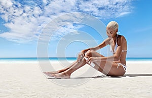 woman on the beach applying sunscreen lotion to legs skin for care and sun protection, in sunny sea day with blue sky. Concept for