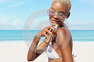 woman at the beach applying sunscreen lotion to skin for care and sun protection, wearing sunglasses in sunny day with blue sky.