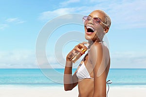 woman at the beach applying sunscreen lotion to skin for care and sun protection, wearing sunglasses in sunny day with blue sky.