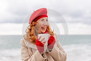 Woman beach in red beret scarf and mitts holding a cup of tea in his hands. Depicting beach relaxation and cozy attire