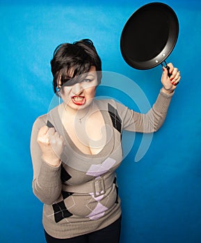 Woman beating with a frying pan on bluw studio background alone