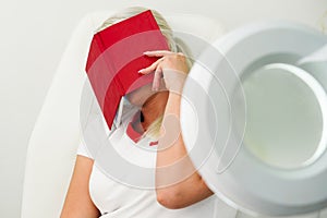 Woman beautician sits on a white armchair, tired, covered her face with a red notebook. Work seven days a week