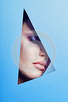 Woman with a beautiful bright makeup and pink lipstick looks through a triangular hole in the blue paper. Advertising