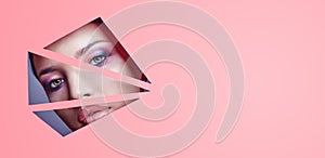 Woman with beautiful bright makeup and pink lipstick looks through triangular slits in pink paper. Advertising cosmetics photo