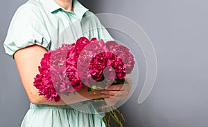 A woman in a beautiful dress holds in her hands a large bouquet of burgundy red peonies on a gray background.