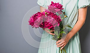 A woman in a beautiful dress holds in her hands a large bouquet of burgundy red peonies on a gray background