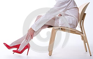 A woman with beautiful legs in white stockings and red shoes sits on a chair on a white