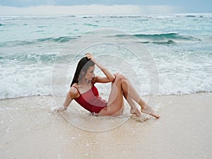 Woman with a beautiful tan tourist in a red swimsuit sitting on the sand on the beach in the ocean in the waves, cloudy