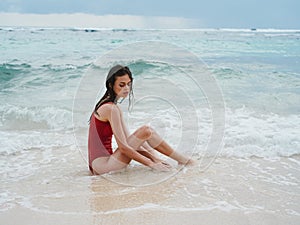 Woman with a beautiful tan tourist in a red swimsuit sitting on the sand on the beach in the ocean in waves, cloudy