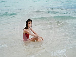 Woman with a beautiful tan tourist in a red swimsuit sitting on the sand on the beach in the ocean in waves, cloudy