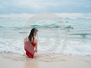 Woman with a beautiful tan tourist in a red swimsuit sitting on the sand on the beach in the ocean in the waves with her