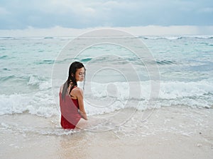 Woman with a beautiful tan tourist in a red swimsuit sitting on the sand on the beach in the ocean in the waves with her