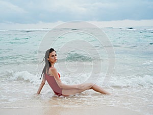 Woman with a beautiful tan tourist in a red swimsuit sitting on the sand on the beach in the ocean in the waves pensive