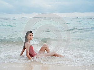 Woman with a beautiful tan tourist in a red swimsuit sitting on the sand on the beach in the ocean in the waves pensive