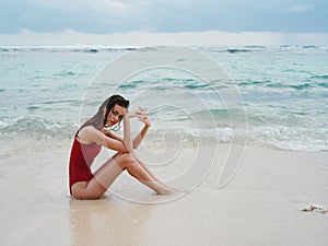 Woman with a beautiful tan tourist in a red swimsuit sitting on the sand on the beach in the ocean in waves, travel and