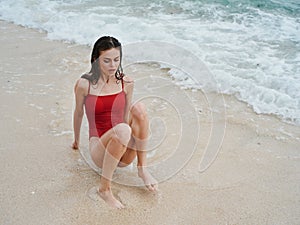 Woman with a beautiful tan tourist in a red swimsuit sitting on the sand on the beach in the ocean in waves, travel and