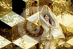 Woman beauty portrait of fashion model in golden dress, waving golden glittering fabric flying in front of cube background