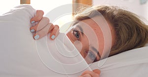 Woman, bed and blanket while peeking, hiding and shy with head on pillow to relax, play and have fun in home bedroom