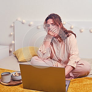 A woman on the bed yawns while sitting at her laptop. The girl works with food on the bed