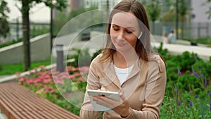 Woman in a beige lingo wearing wireless headphones uses a tablet while sitting on a bench in the park