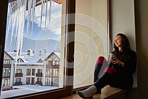 Woman being cold sits on windowsill, warming up with tea mug, looking at snow covered mountains. Female drinks coffee in
