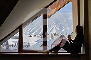 Woman being cold sits on windowsill, warming up with tea mug, looking at snow covered mountains. Female drinks coffee in