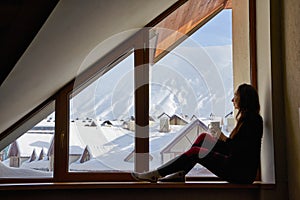 Woman being cold sits on windowsill, warming up with tea mug, looking at snow covered mountains. Female drinks coffee in