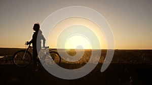 A woman with a bicycle walks along the top of a hill against the background of a bright sunset. Silhouette. 4K.