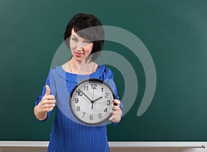 Woman with big clock shows Okey gesture, chalk board background, time and education concept, green background, studio shot