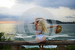 Woman in bikini with a beautiful physique sitting on beach looking at ocean view on sunset in swimwear. Slim figure girl