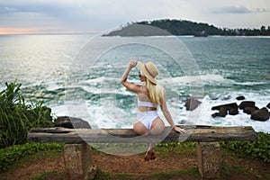 Woman in bikini with a beautiful physique sitting on beach looking at ocean view on sunset in swimwear. Slim figure girl