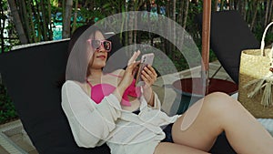 Woman in bikini lying on the deck chair and using smartphone near the pool